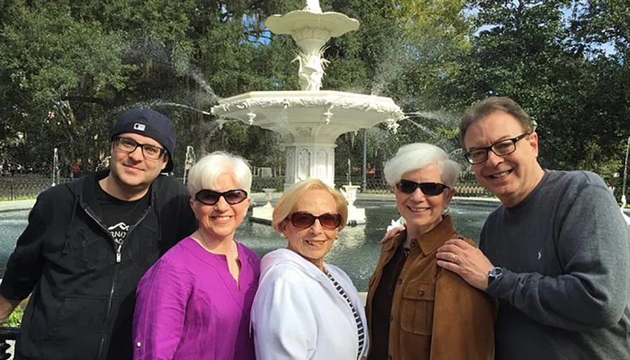 Five adults are smiling in front of an ornate fountain in a sunny park setting.