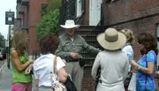 A tour guide in a wide-brimmed hat is explaining something to a group of attentive women on a city street.