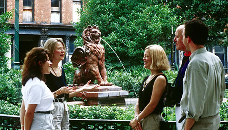 Four individuals are engaged in a conversation in front of a bronze statue of a lion in a park-like setting.