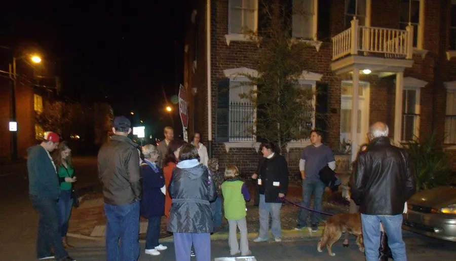 A group of people, some attentively looking towards one person, is gathered on a street at night, possibly for an event or tour, with a dog also present.