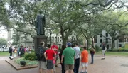 The image depicts a group of people gathered around a tour guide in a park with a statue and large trees in the background.