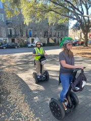 Two people, one wearing a helmet with a quirky alligator design and the other sporting a vest, are riding Segways on a tree-lined path with historic buildings in the background.