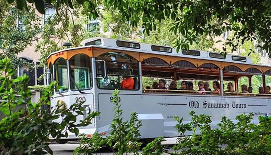 A trolley bus filled with tourists is navigating through a lush, tree-lined street as part of an Old Savannah Tours sightseeing experience.