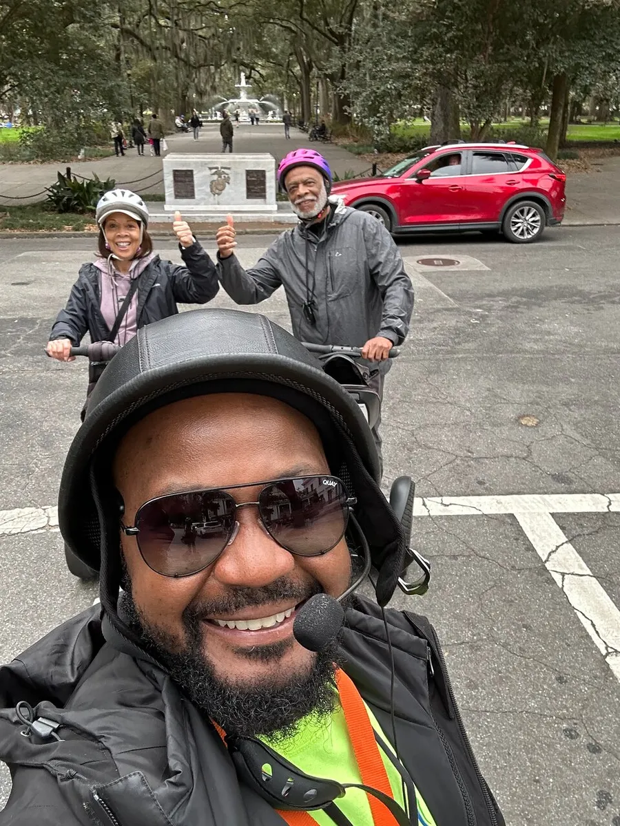 Three people wearing helmets and casual attire are smiling for a selfie on a tree-lined street, with two giving a thumbs-up and a fountain visible in the background.