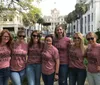 A group of seven smiling women wearing matching pink t-shirts that read SIDE PARTS  SKINNY JEANS pose together outdoors