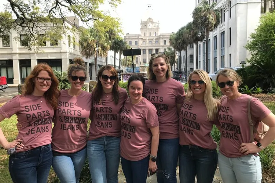 A group of seven smiling women wearing matching pink t-shirts that read SIDE PARTS & SKINNY JEANS pose together outdoors.