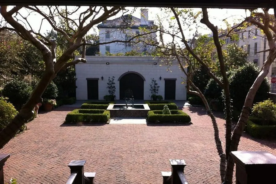 The image shows a serene courtyard with symmetrical hedge patterns, a central fountain, and brick paving, flanked by a white building with an arched doorway and framed by leaf-bare trees under a bright sky.