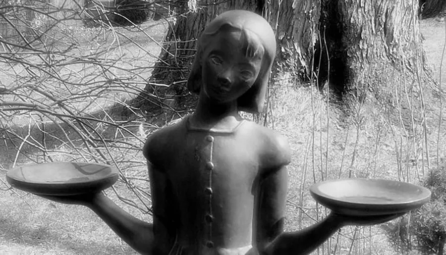 The image shows a black and white photo of a statue of a girl with outstretched arms, each holding a plate, set against a natural backdrop.