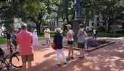 A group of people is standing outdoors in a park or square, likely listening to a guide or speaker near a statue and a flowerbed.