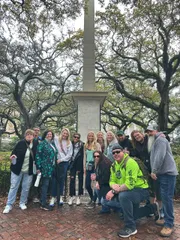 A diverse group of people posing for a photo in a park with a monument and sprawling oak trees in the background.