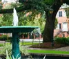 This image shows a green water fountain with water cascading down its tiers surrounded by lush greenery with a lamp post and a red brick building in the background suggesting an urban park setting