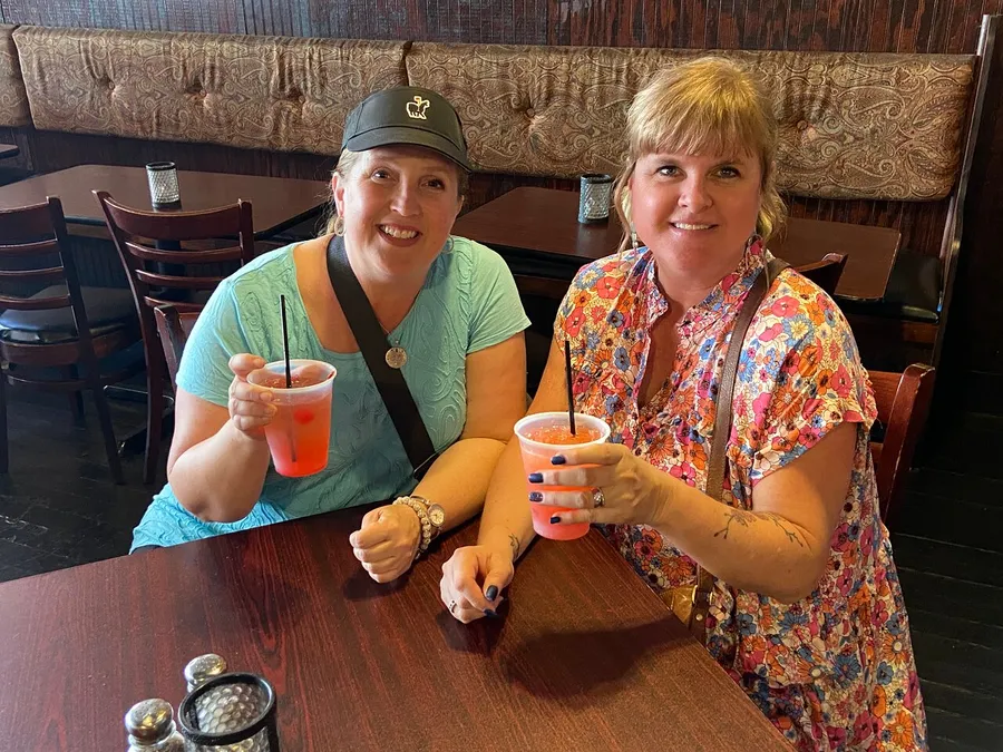 Two smiling women are sitting at a restaurant table, each holding a cup of a pink-colored beverage, seemingly enjoying each other's company.