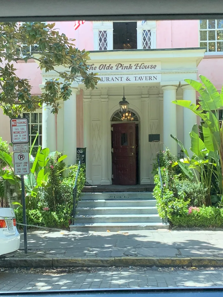 The image shows the entrance to The Olde Pink House Restaurant & Tavern, a pink-colored historic building with white trim, a red door, and greenery flanking the steps.