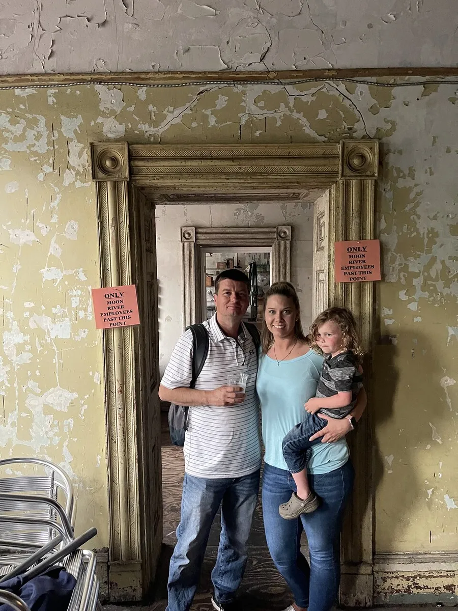 Three people are standing together smiling in front of a peeling wall inside a room with an ornate mirror and signs that say ONLY EMPLOYEES PAST THIS POINT.