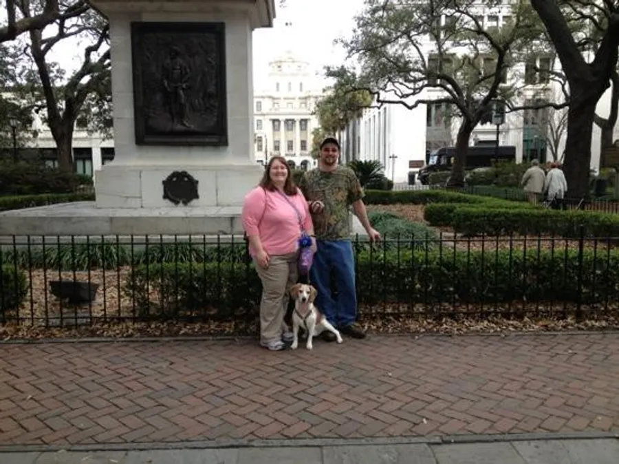 A man and a woman pose for a photo with their dog in what appears to be a park with a statue and a building in the background.