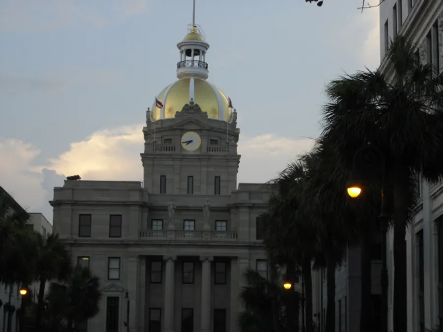 The image features a majestic building with a gold-domed clock tower, flanked by palm trees and street lamps against an evening sky.