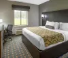 The image shows a neatly arranged hotel room with a queen-sized bed a work desk and a patterned carpet