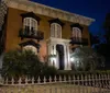 An elegant historic building is illuminated at night showcasing its ornate wrought-iron balcony and fencing with lush greenery adding to its stately charm