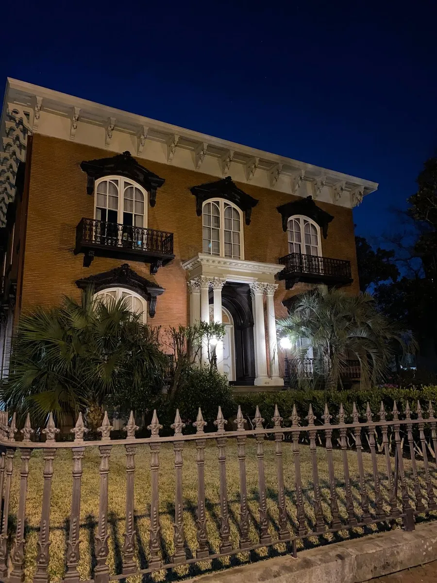 An elegant historic building is illuminated at night, showcasing its ornate wrought-iron balcony and fencing, with lush greenery adding to its stately charm.