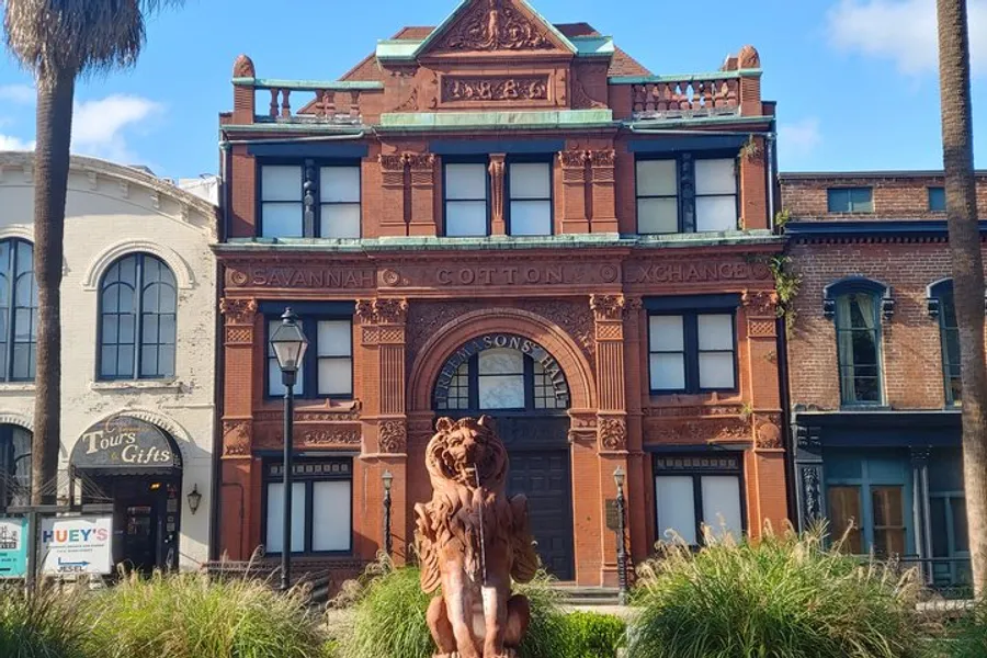 The image displays the front view of the ornate Savannah Cotton Exchange building, distinguished by its red brick facade and historical architecture, accompanied by a lion statue in the foreground.
