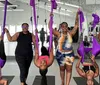 A group of smiling people are using purple aerial silks in a fitness studio with one person doing an upside-down pose