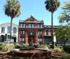 The image features a picturesque urban square with a lion sculpture fountain in the foreground and a historic red-brick building flanked by palm trees under a clear blue sky