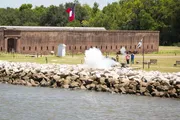 This image shows a historic fort with a large Texas flag flying, people nearby, and a cannon firing, creating a large cloud of smoke along the rocky shoreline.
