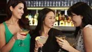 Three women are smiling and toasting with cocktails at a bar.