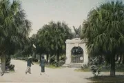 Two individuals are meandering down a palm-lined pathway toward a white stone archway in what appears to be an old, hand-colored photograph.