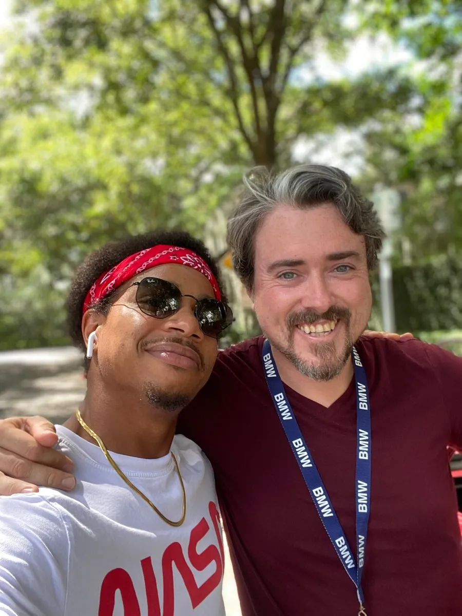 Two men are smiling for a selfie outdoors, with one wearing sunglasses and a red headband, and the other donning a burgundy shirt and a lanyard.