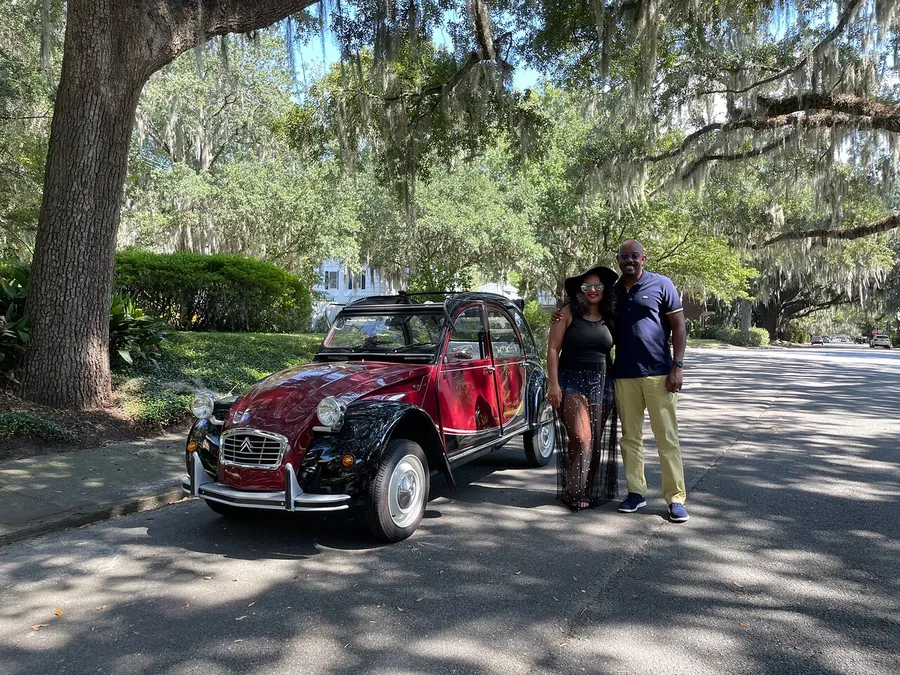 A smiling couple stands beside a classic red car under the shade of large trees draped with Spanish moss.
