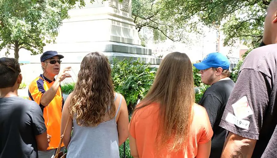 A tour guide in an orange vest is explaining something to a group of attentive listeners outdoors.