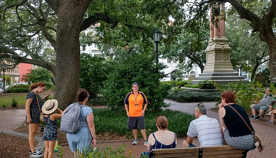 A group of people is gathering around a tour guide in an orange shirt in a park with lush trees and a monument.