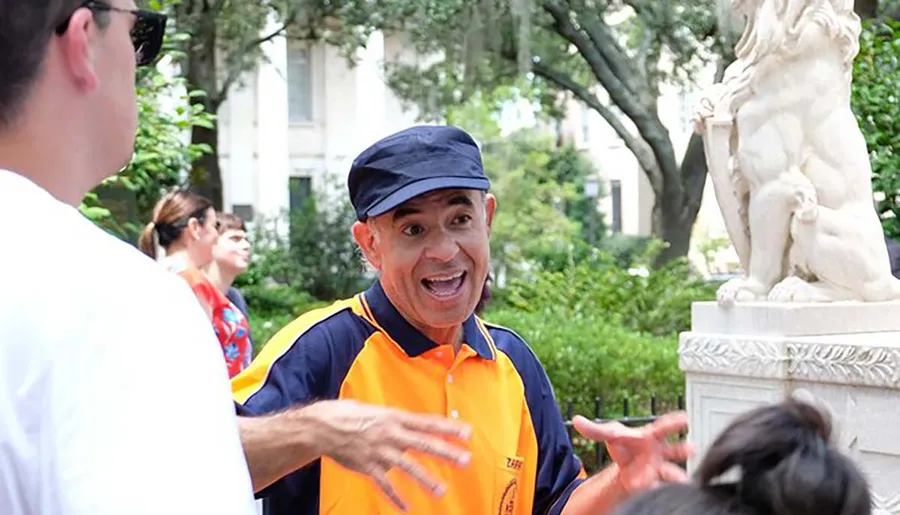 A man in an orange shirt and cap enthusiastically gestures as he speaks to a group of people in a park with a statue in the background.