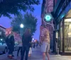People are casually interacting on a lively city street during dusk with shops and streetlights surrounding them