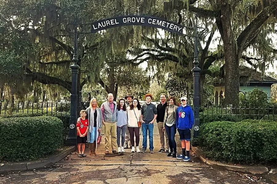A group of people is posing for a photo in front of the entrance to Laurel Grove Cemetery, which is adorned with Spanish moss-draped trees.