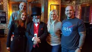 The image shows five people smiling for a group photo, with one man in a top hat and red tie standing in the center.
