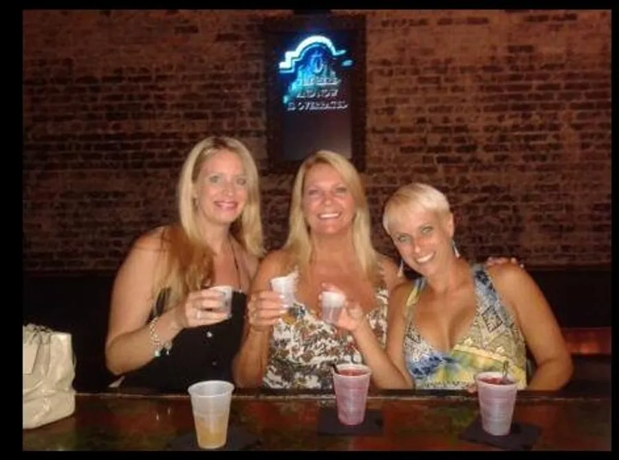 Three individuals are smiling for the camera while sitting at a bar, holding drinks, with a neon sign in the background.