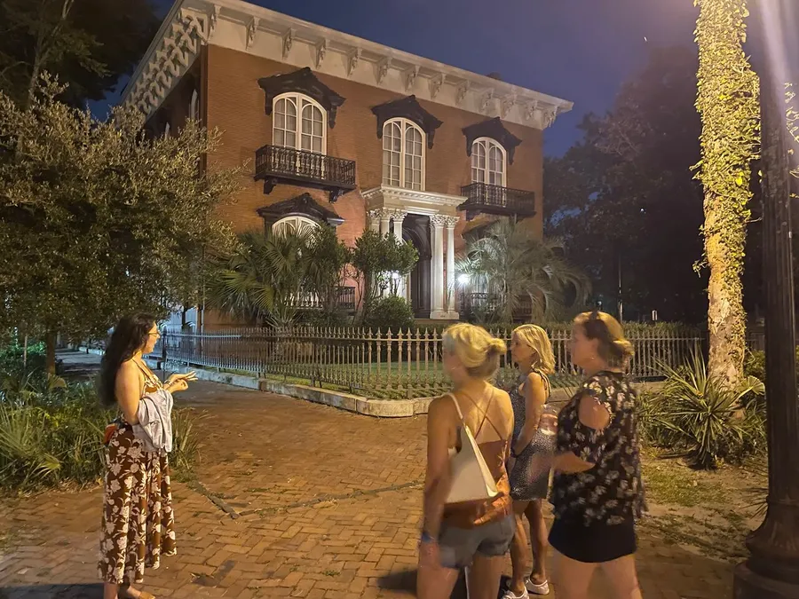 A group of people stands near a wrought iron fence at night, with one person pointing at a stately brick building adorned with white columns and balconies, surrounded by lush greenery.