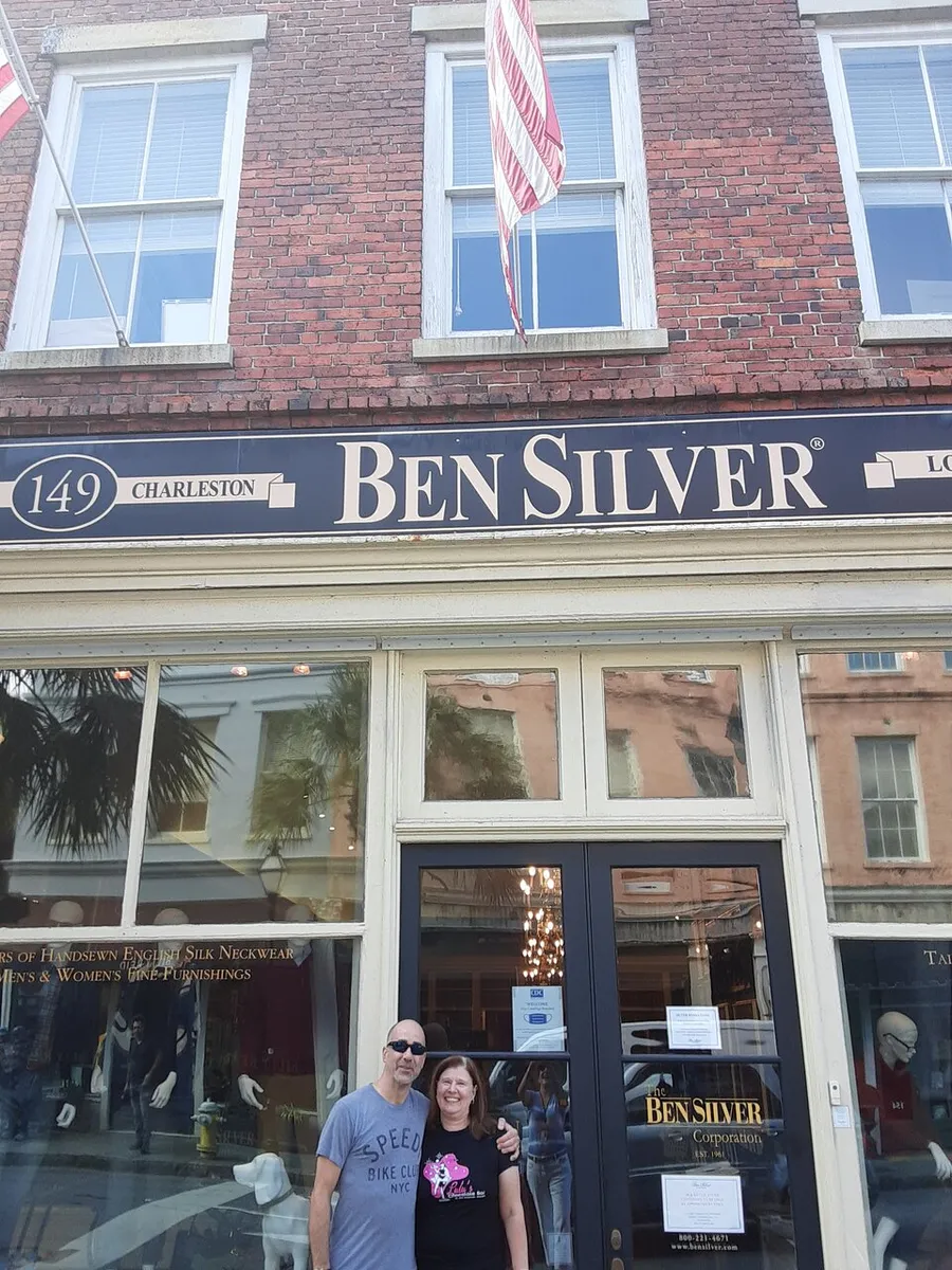 A man and a woman stand smiling in front of the Ben Silver storefront on a sunny day, with American flags hanging above them.
