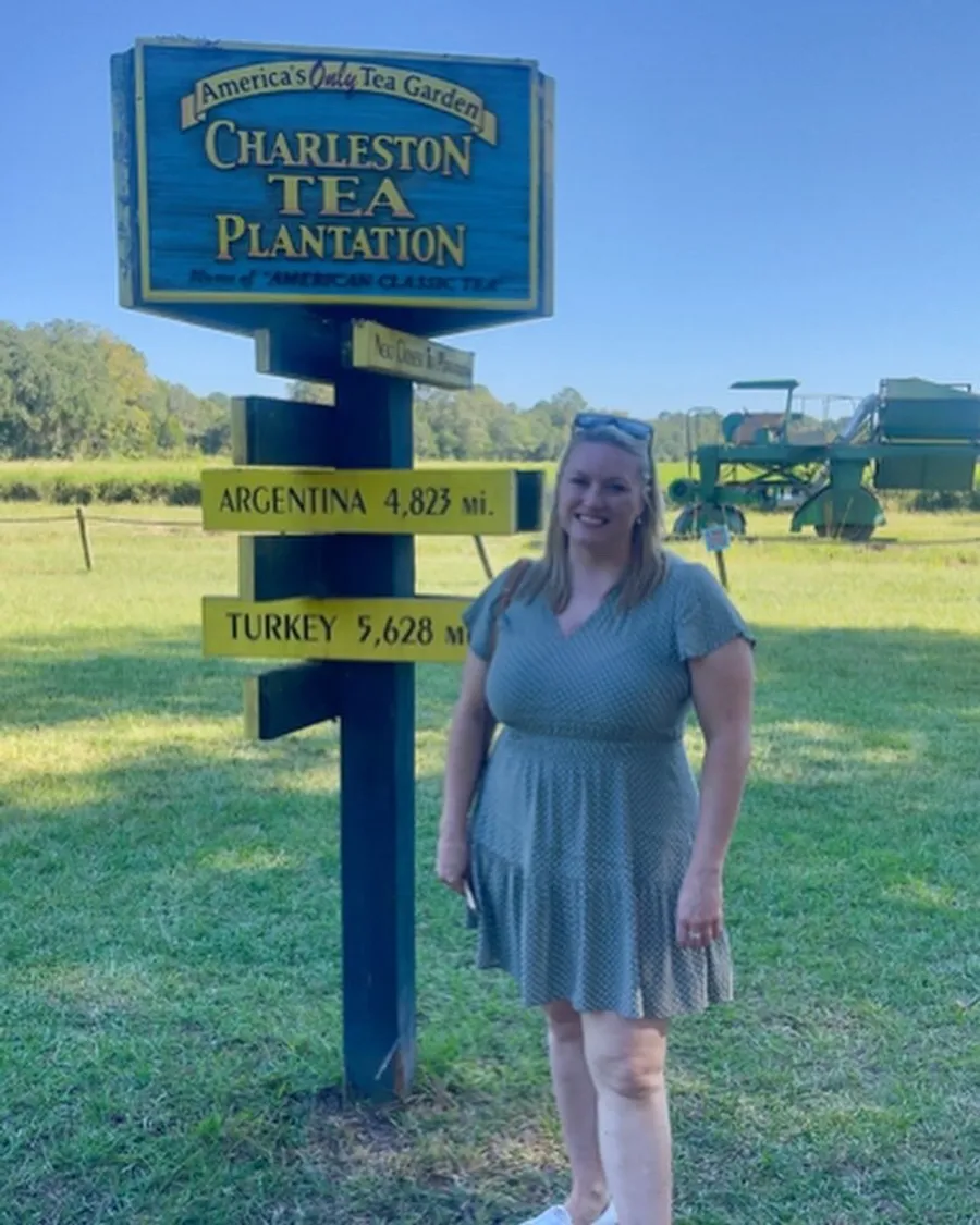 A woman in a dress is standing and smiling next to a blue and yellow sign that reads Charleston Tea Plantation with directional arrows pointing to Argentina and Turkey noting their distances in miles.