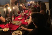 Guests are gathered around a festively set dinner table with lit candles, red accents, and elegant tableware, participating in a dimly lit, intimate gathering.