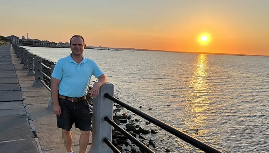 A man is smiling at the camera while standing by a seaside railing during sunset.