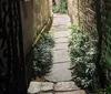 The image shows a narrow overgrown pathway between old brick and plaster walls with a glimpse of a wrought iron gate on the left creating an atmosphere that is both quaint and slightly mysterious