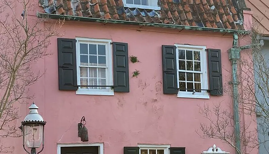 The image shows the upper facade of a charming pink house with black shutters, terracotta roof tiles, and a classic street lamp to the side.
