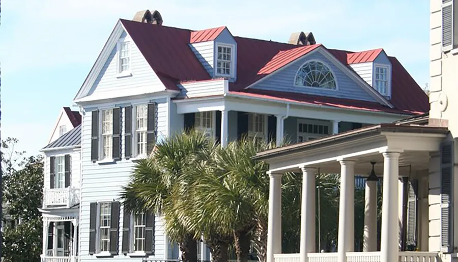 The image depicts a large, two-story white house with a red roof, traditional shutters on the windows, a second-story balcony, and a front porch supported by columns, all set against a clear blue sky and complemented by a palm tree in the foreground.