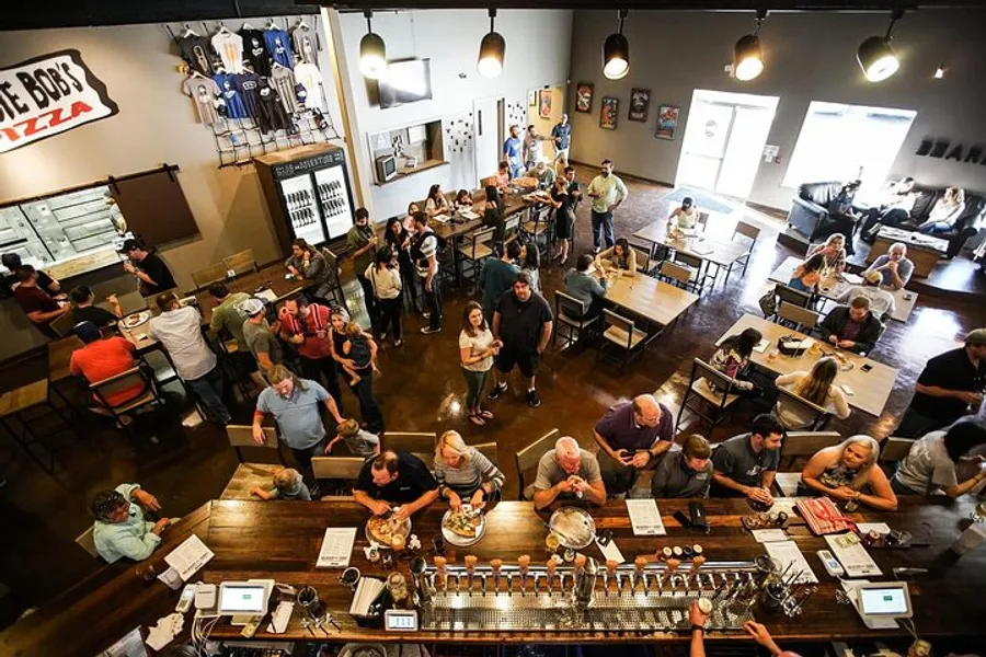 A bustling brewery or pub with patrons engaged in eating, drinking, and socializing, featuring a bar area, dining tables, and a pizza service counter.