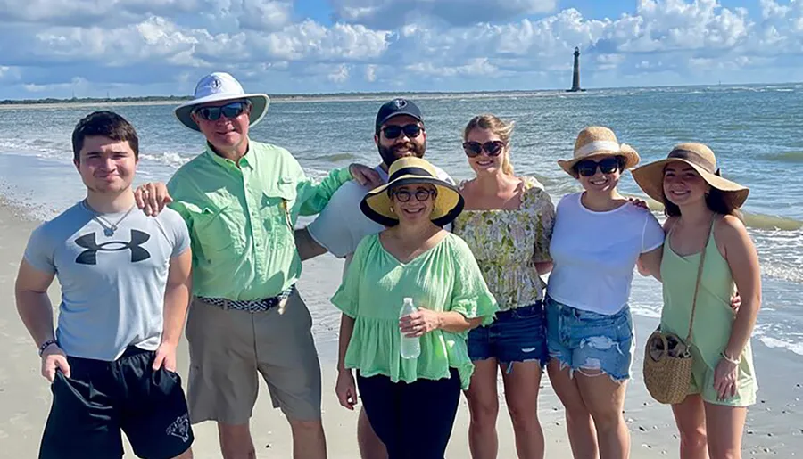 A group of people poses smiling on a sunny beach with a lighthouse visible in the distance.