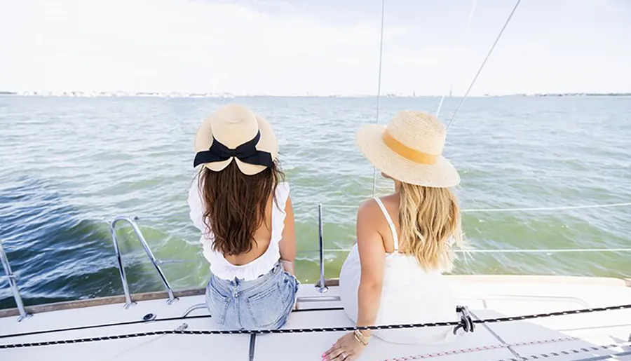 Two people are seated at the stern of a sailboat, looking out at the water while wearing stylish hats.