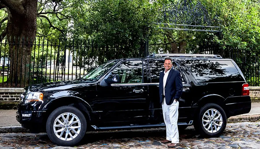 A smiling man in business attire is standing next to a black SUV parked in front of a wrought iron fence and trees.